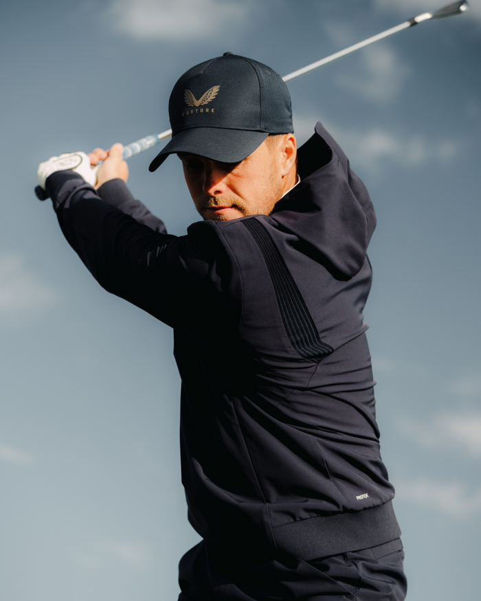 Golfer in midnight navy performance hoodie mid swing with a club against a cloudy sky.