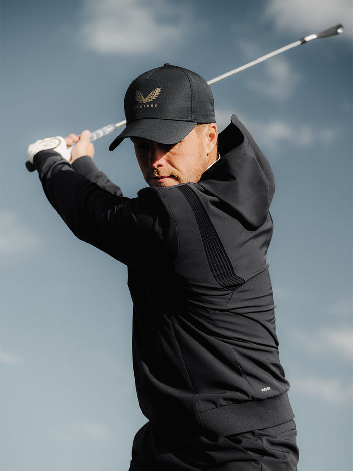 Golfer in black Pinnacle Performance Hoodie mid swing with a golf club and Castore logo on the chest