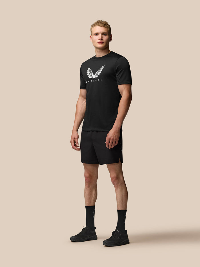 Full-body side view of a man in black Adapt Training T-Shirt and shorts with white Castore wings logo on chest