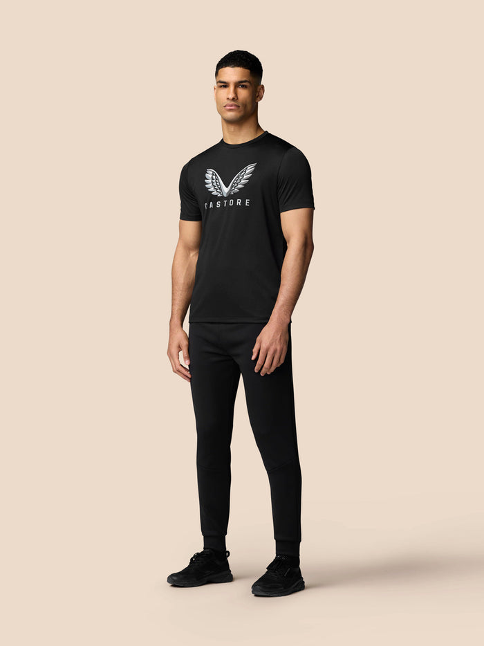 Male model wearing black graphic T shirt with silver wing logo on chest and black joggers.