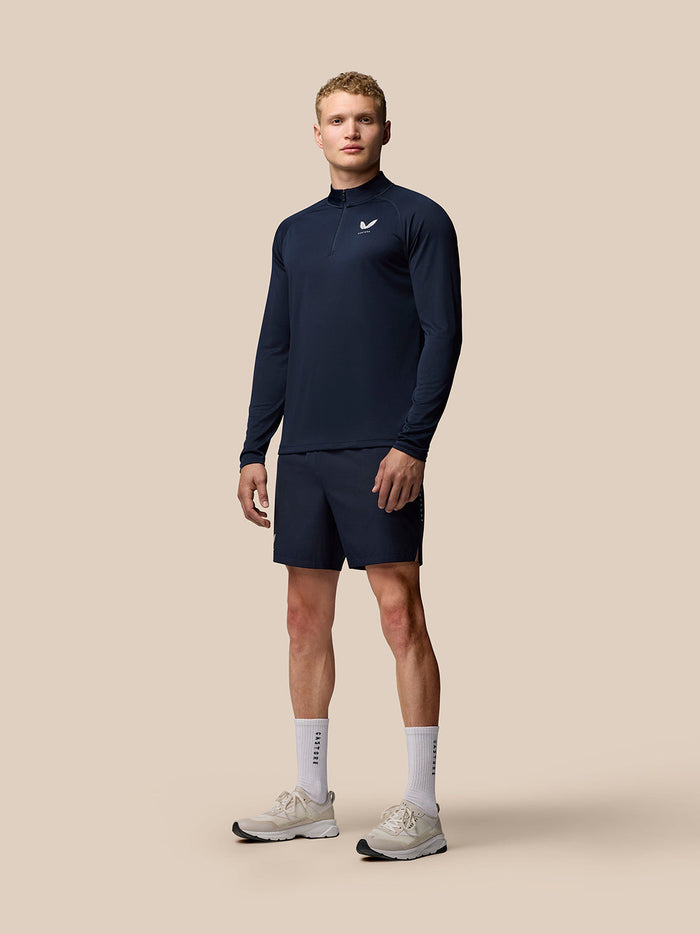 Full body shot of male model in navy long sleeve Adapt Quarter Zip Top with logo on left chest, paired with navy shorts and white sneakers.