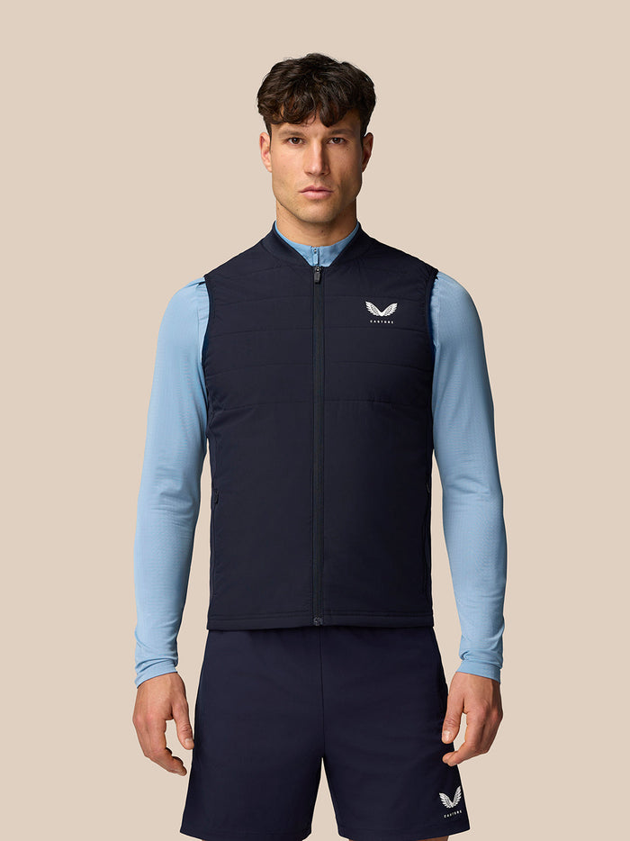 Male model wearing navy Apex Training Gilet over a light blue long sleeve top in a full body shot.