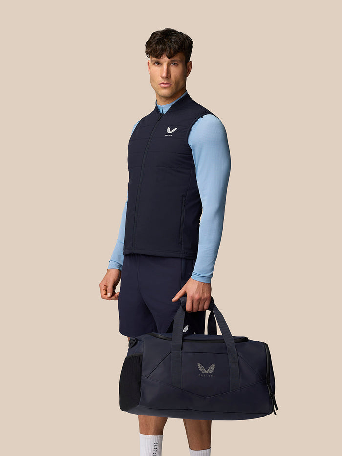Male model wearing navy Apex Training Gilet over a light blue top, carrying a navy Castore duffle bag.