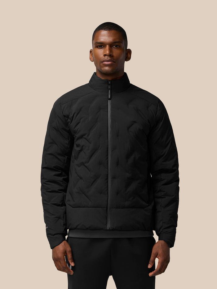 Male model wearing a black padded jacket with stand collar and full zip against a beige background.