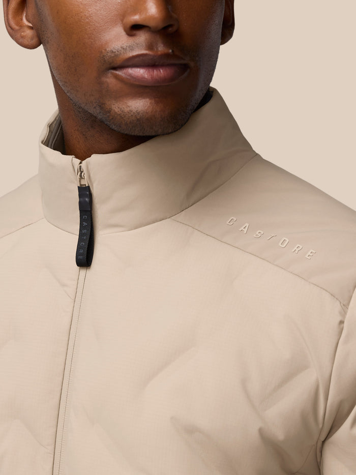 Close up of a man wearing mushroom beige padded jacket with a zip collar and CASTORE branding on the chest.