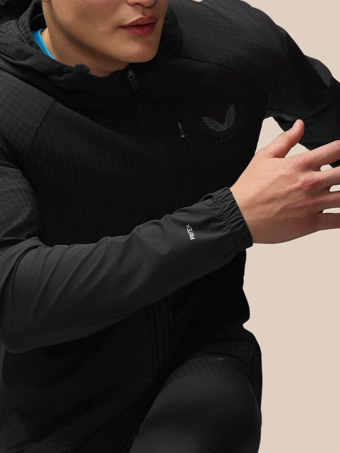 Full-body front view of model in black Flex Winterised Jacket with hood against a beige background.