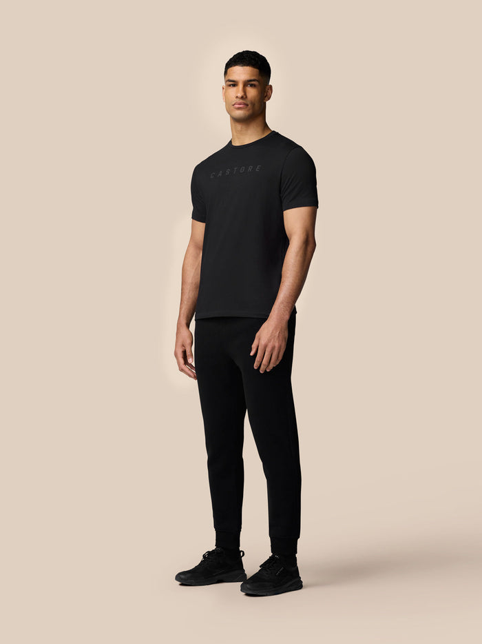 Full body front view of man in black Castore graphic T shirt with black joggers and sneakers against a beige background