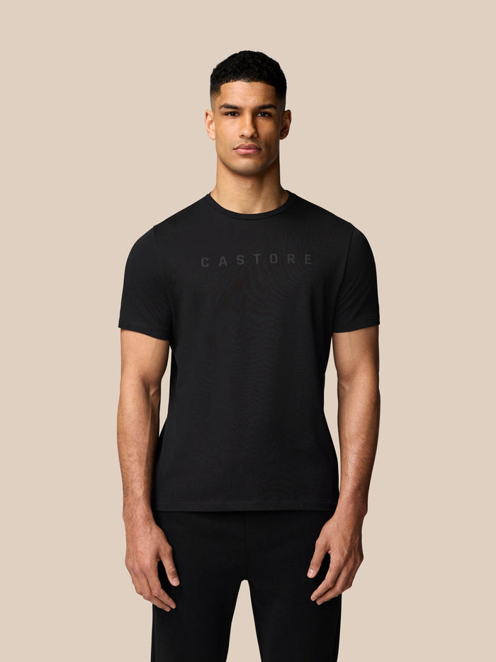 Front view of man wearing black Castore graphic T shirt with subtle chest logo on beige backdrop