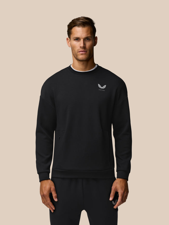 Front view of a man in a black crew neck Travel Lifestyle Sweatshirt with a white chest logo against a beige background
