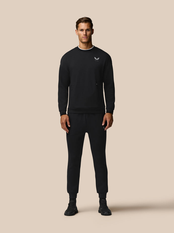 Front view of a man wearing a black crew neck travel lifestyle sweatshirt with a left chest logo against a beige backdrop