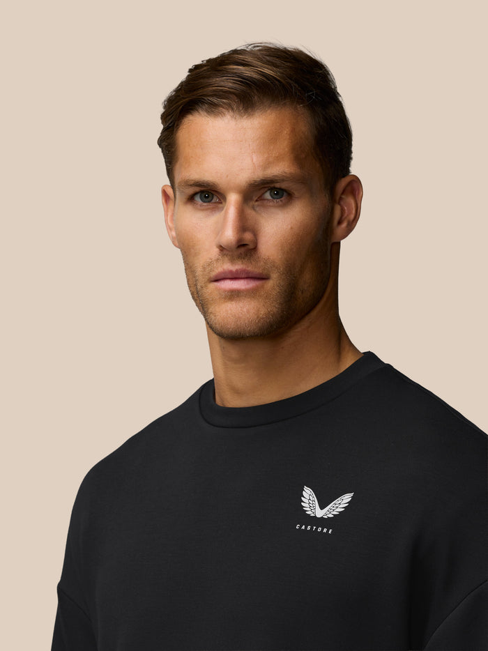 Portrait of a man in a black crew neck sweatshirt with a white wing logo on the left chest against a beige backdrop