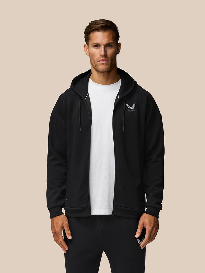 Man wearing black travel zip hoodie over a white t shirt with left chest Castore logo, standing against beige background.