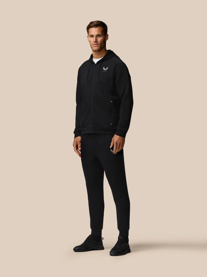 Full body shot of a man in black travel zip hoodie and black joggers, Castore logos on chest and leg, beige background.