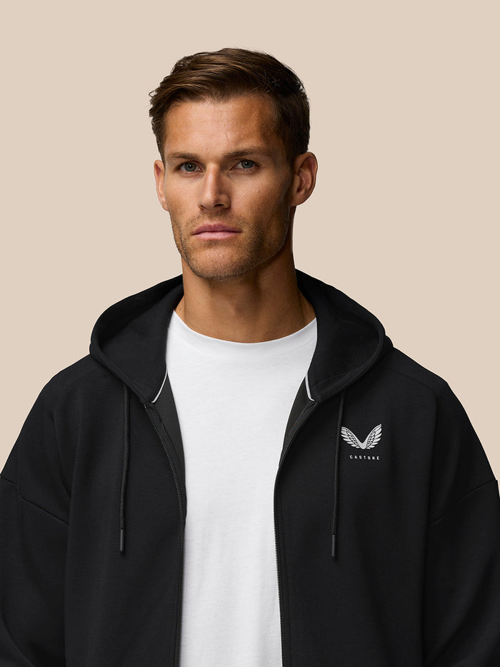 Close up of a man wearing the black travel zip hoodie with left chest Castore logo visible against a beige backdrop.