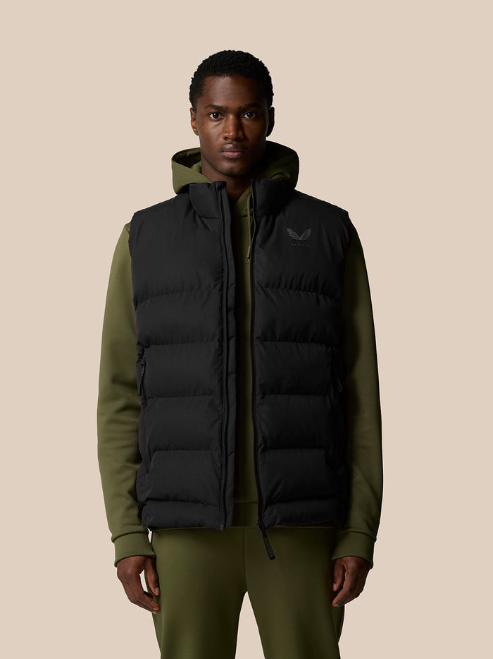 Male model wearing a black sleeveless puffer vest over an olive hoodie, front zip and chest logo visible