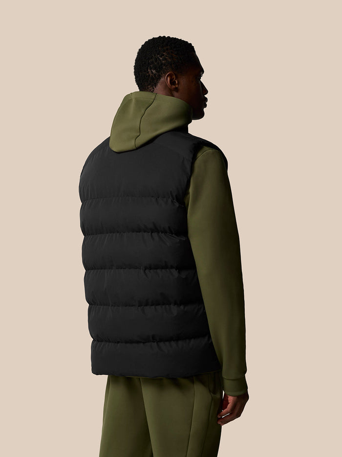 Back view of a man wearing a black sleeveless puffer gilet over an olive green hoodie