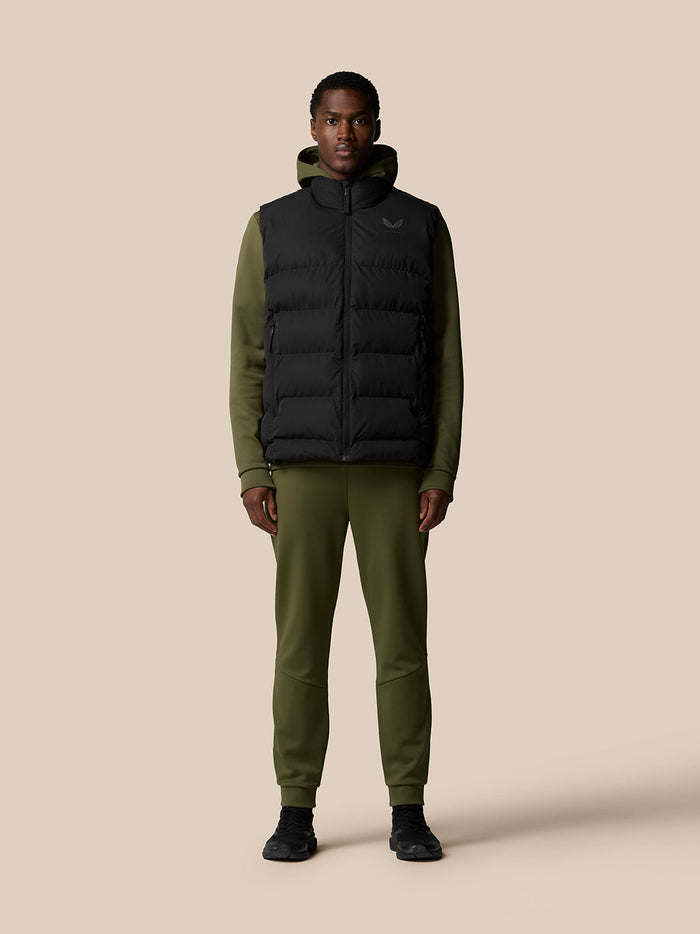 Front view of a man wearing a black puffer gilet over an olive green hoodie standing against a beige background