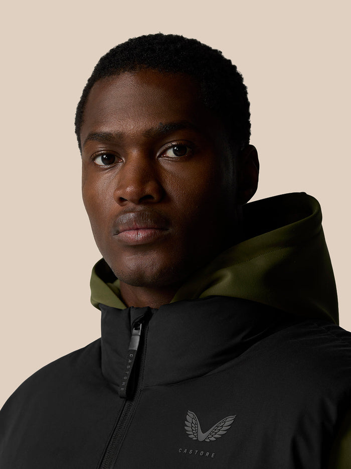 Portrait of a man wearing a black ProTek Puffer Gilet with olive hood lining against a beige background