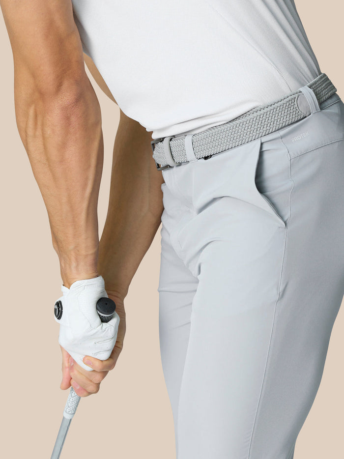 Side view of a man wearing light grey golf trousers with a woven belt, white polo, gripping a golf club.