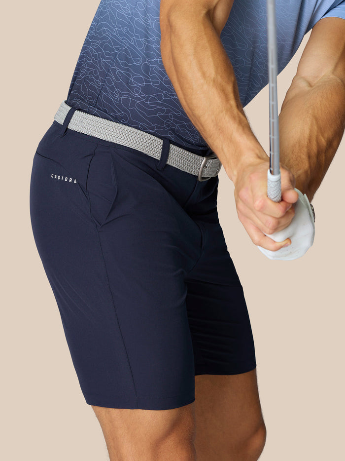 Male model wearing navy golf shorts with a light gray braided belt; Castore logo on the thigh and ProTek branding at the waistband.