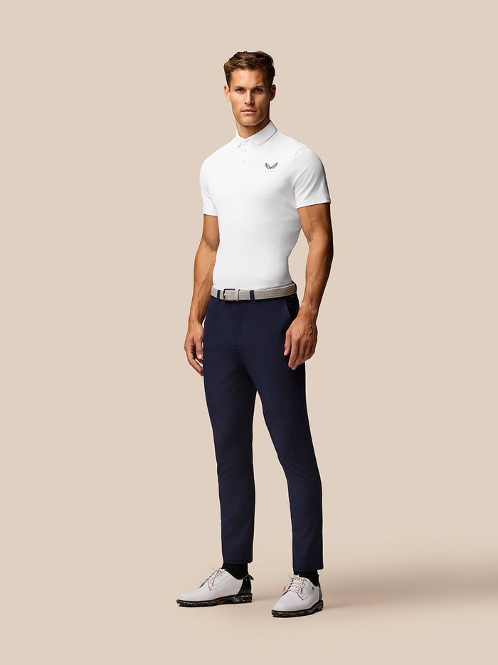 Full body shot of a man in white Golf Club Essential Polo with navy trousers and a woven belt against a beige background
