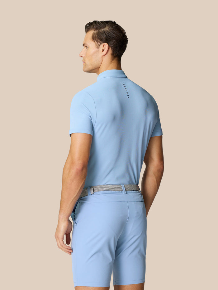 Back view of a man in sky blue golf polo with Castore branding along the upper back against a beige backdrop