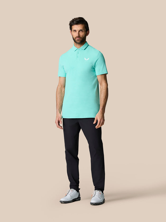 Full body shot of a man in a light aqua Golf Club Essential Polo with a white Castore logo on the chest, paired with dark trousers and light sneakers.
