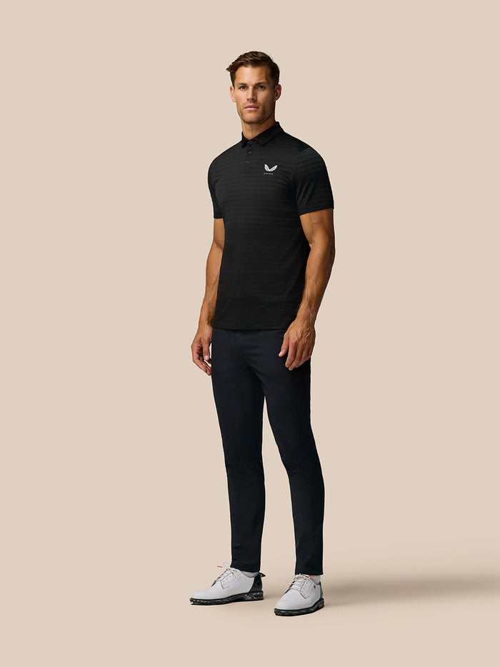 Full-length view of a man in a black Pinnacle Polo with Castore logo, dark pants and white golf shoes.