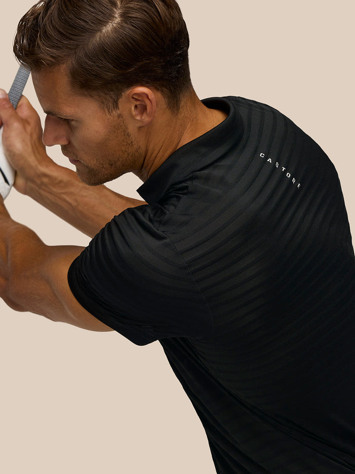 Close-up of a golfer in the black Pinnacle polo mid swing gripping a club with Castore branding visible on the back