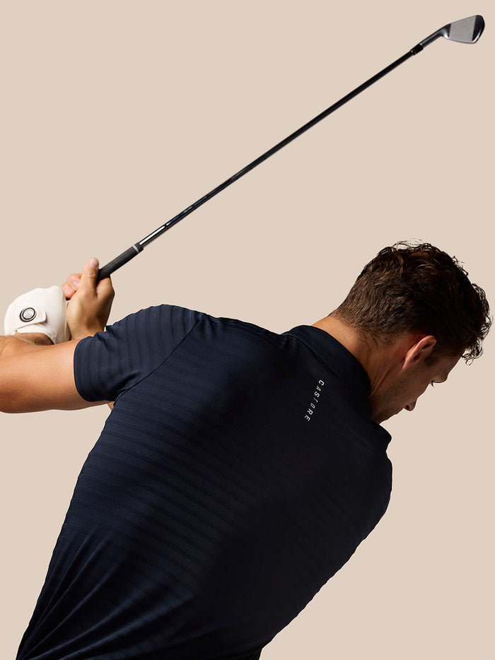 Golfer wearing navy Pinnacle Pique Stripe Polo mid swing against a beige background