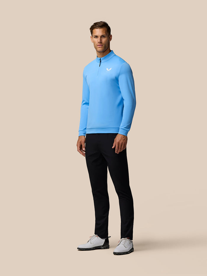 Full-body shot of a male model in a cool blue quarter zip golf top with left chest logo, black pants and white shoes