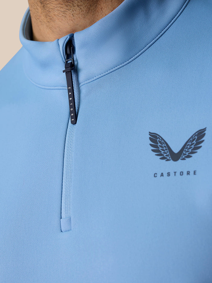 Close-up of the sky blue top at the collar with the zipper and Castore logo with wings on the chest.