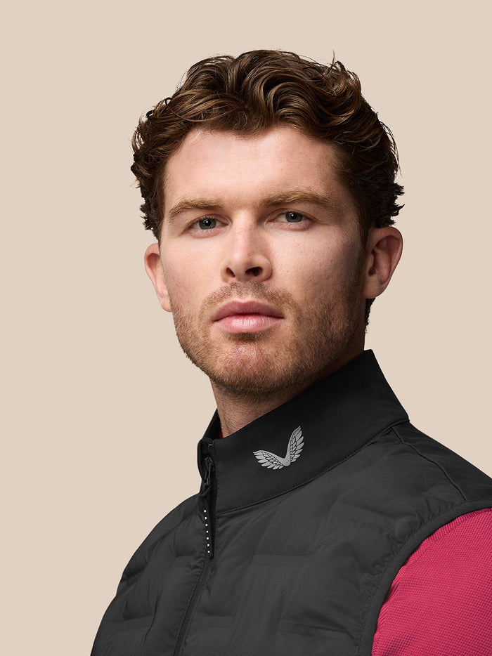 Three quarter back view of a man wearing a black Golf Pinnacle Gilet with branding on the collar and a pink sleeve visible.