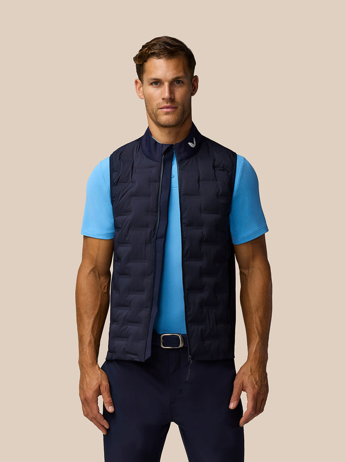 Male model wearing midnight navy Golf Pinnacle Gilet over a light blue polo with front zip and stand collar