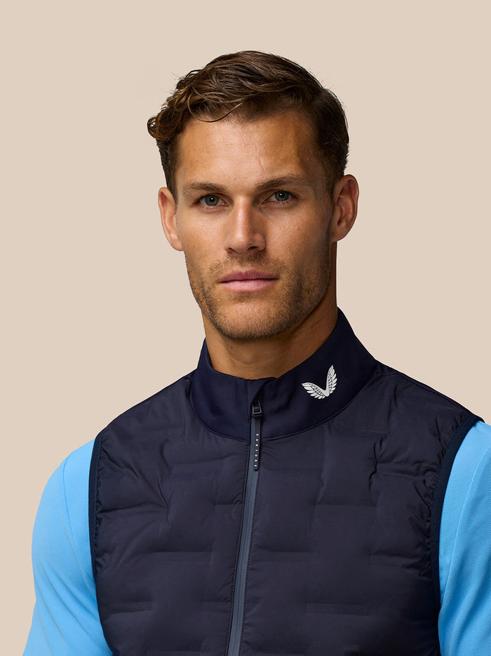Male model wearing midnight navy golf gilet with stand collar over a light blue long sleeve top