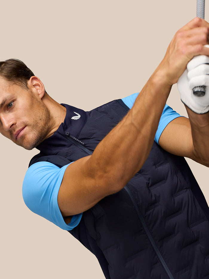 Male golfer wearing midnight navy Pinnacle Gilet over a light blue shirt, in a mid swing pose.