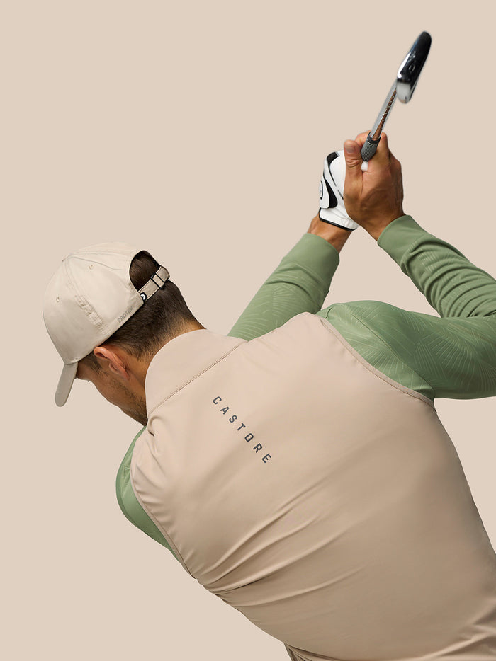 Golfer in mushroom Pinnacle Gilet mid swing, showing the quilted front panel and neutral color.