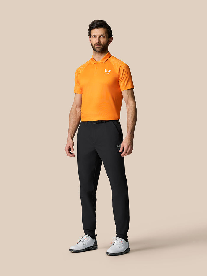 Male model wearing black Golf Pinnacle 32 trousers with an orange polo on a beige studio backdrop