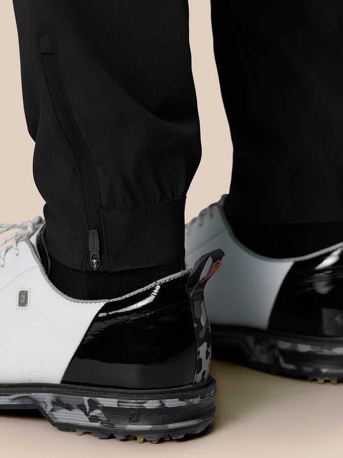 Close-up of black golf trousers cuff with lower leg zipper and elastic cuff, white sneaker visible.