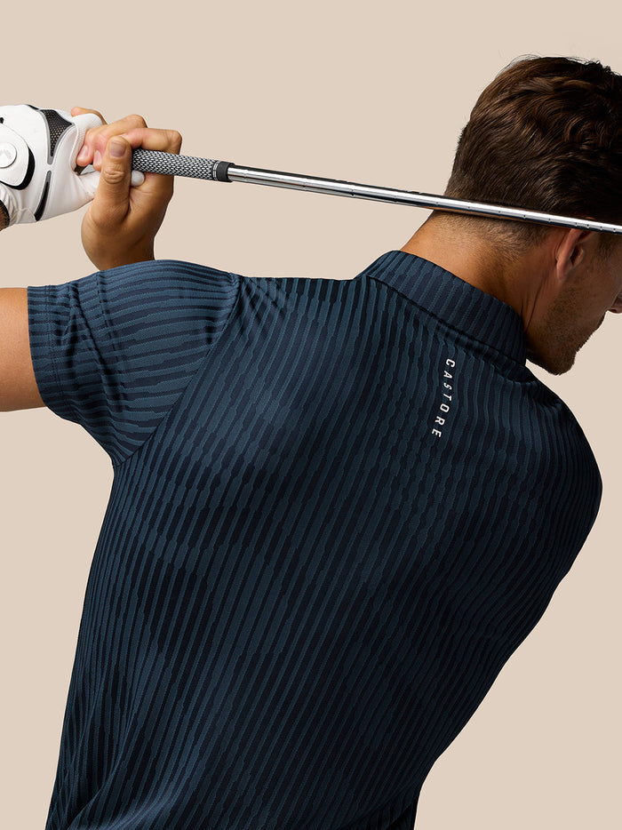 Back view of golfer in midnight navy Golf Pinnacle Glow Polo with striped knit and Castore logo along the back during a swing.