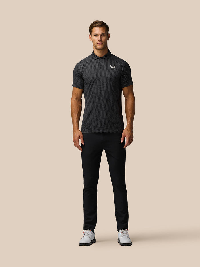 Full body view of a man wearing Golf Club Glow Printed Polo in gunmetal grey with black pants against a beige background