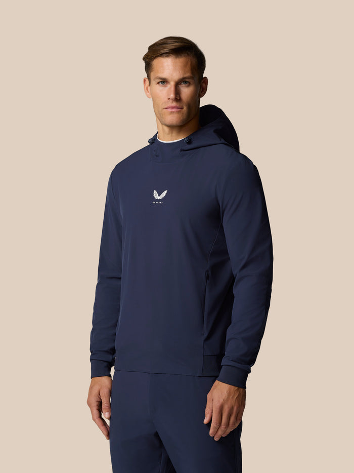 Male model wearing navy Castore Golf Pinnacle Performance Hoodie with chest logo against a beige background